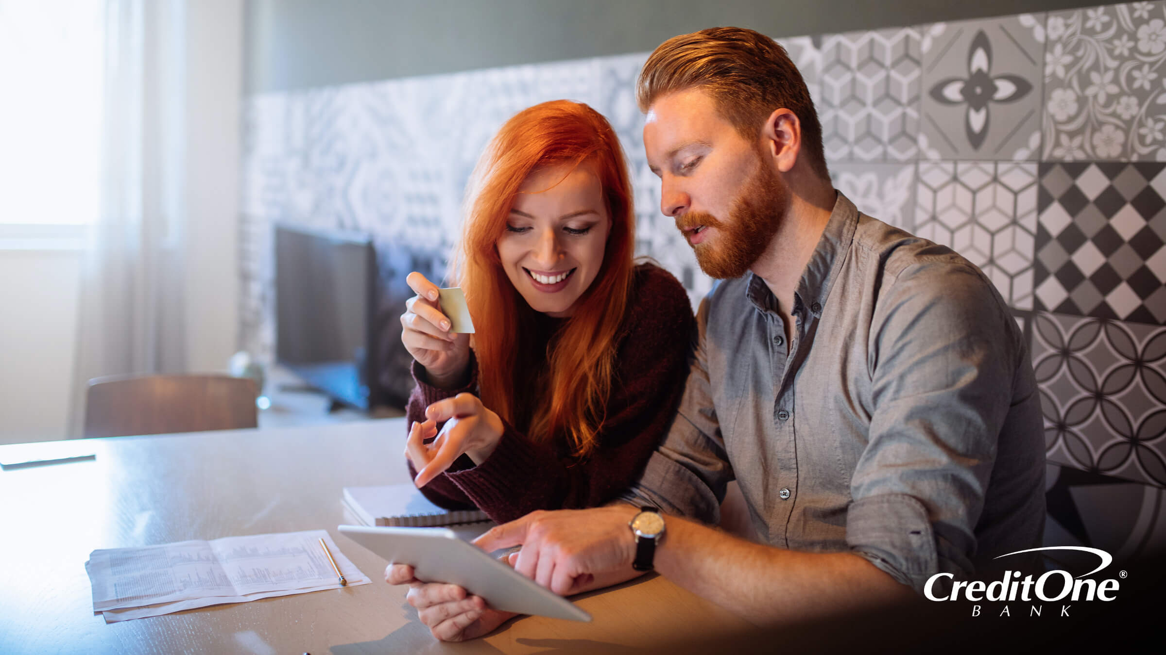 A couple sits at the kitchen table using a tablet while looking at paperwork and holding a credit card, indicating that one of them may be an authorized user.