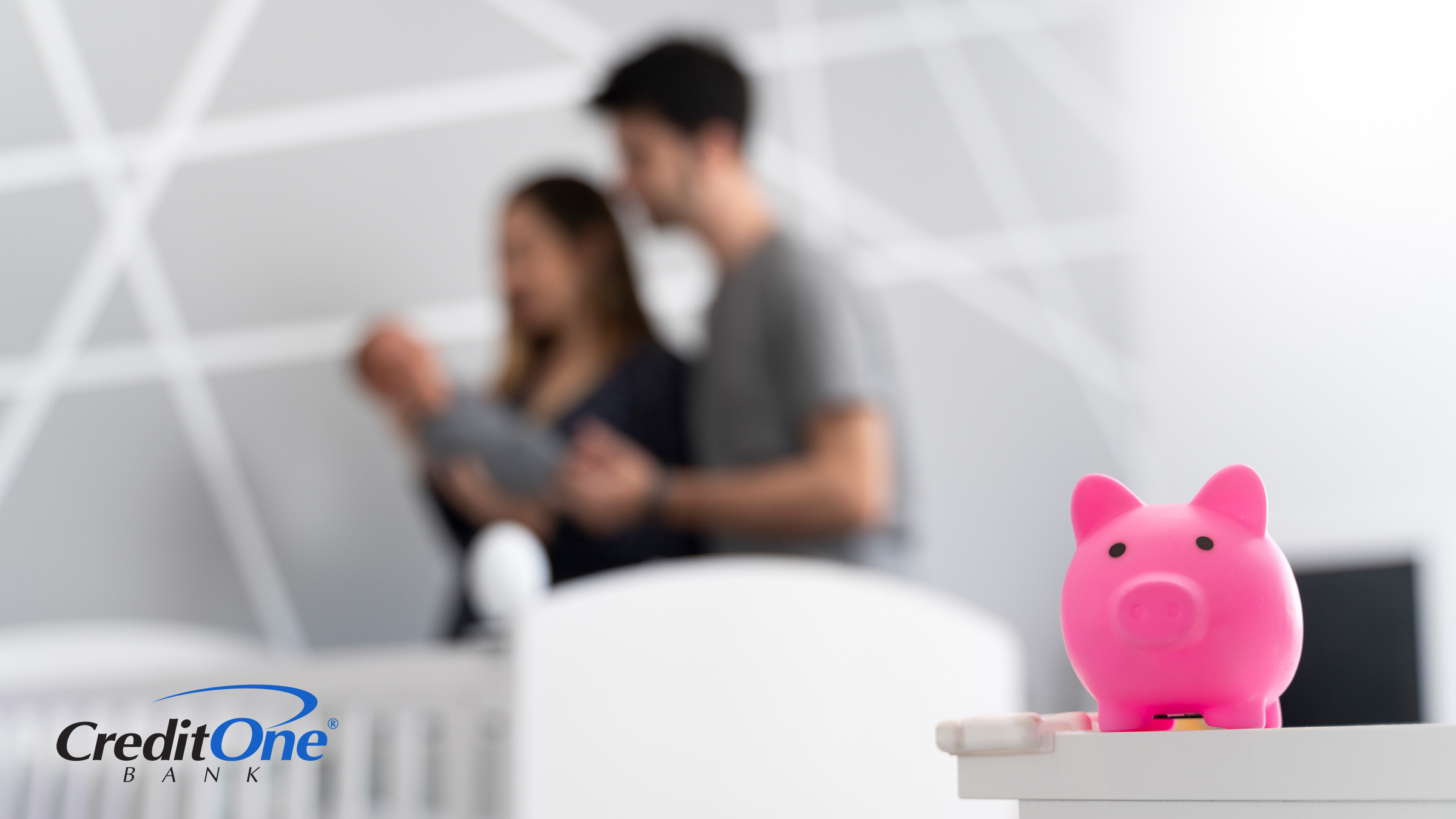 A piggy bank rests on a dresser in the foreground while, just out of focus in the background, a couple places their baby in its crib, symbolizing the ever-present costs of raising a child.
