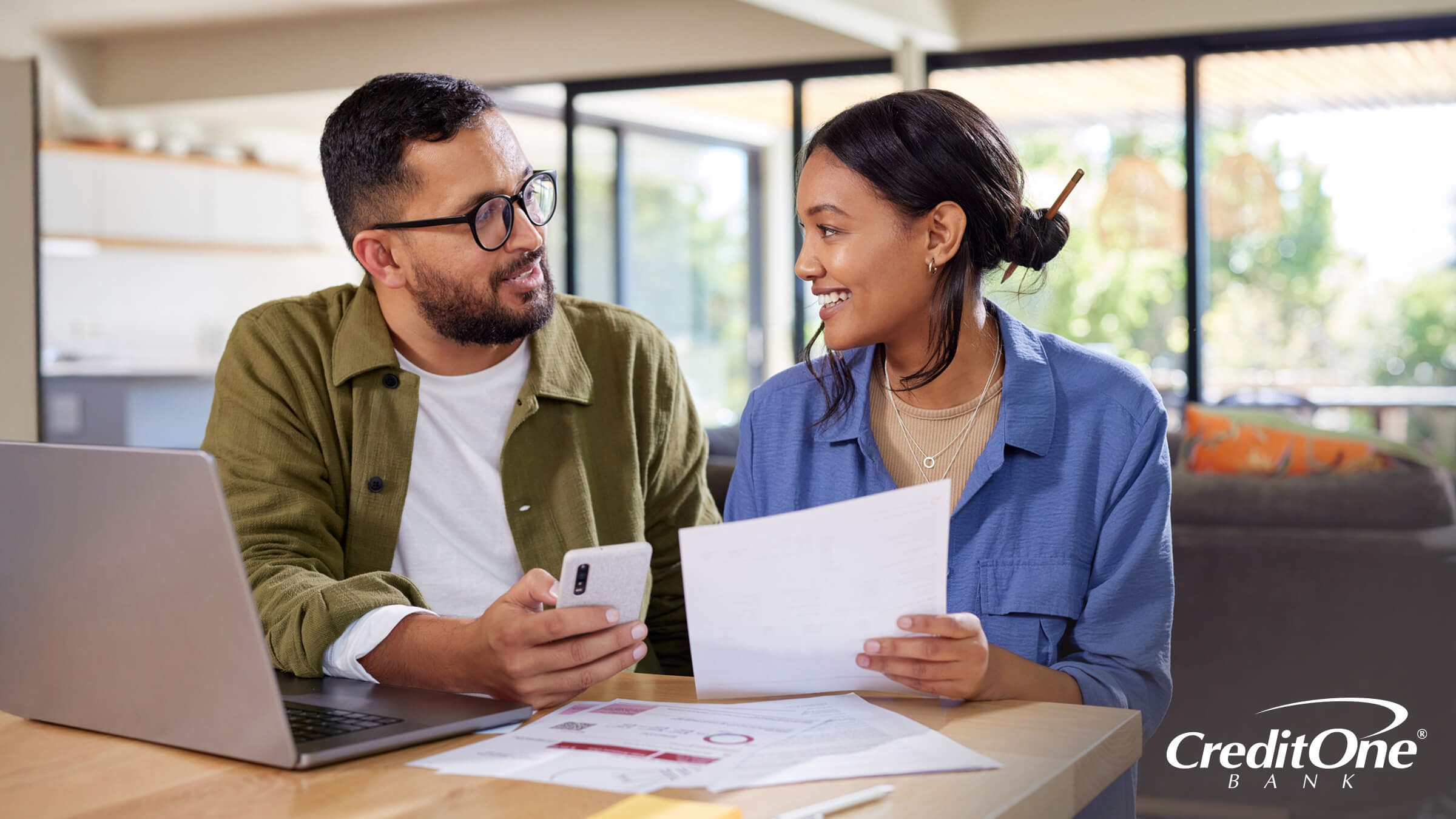A couple sits together at a table, which has financial paperwork and a laptop on it. They appear to have just come to an agreement, perhaps adopting a budgeting system like the 70/20/10 rule.