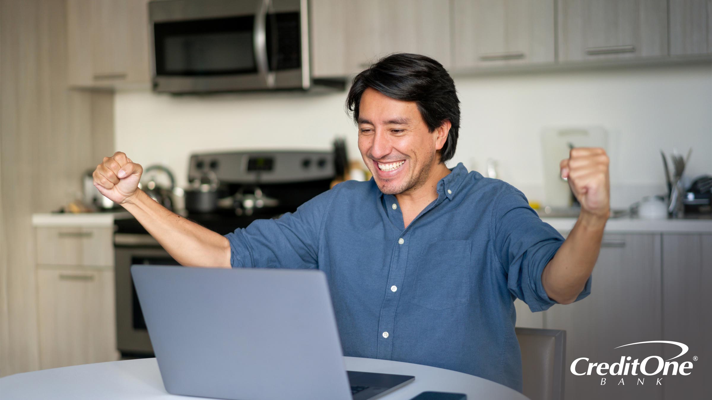 A man sitting at the kitchen table with his laptop smiles broadly and puts both fists in the air as if he’s celebrating his improved credit score.