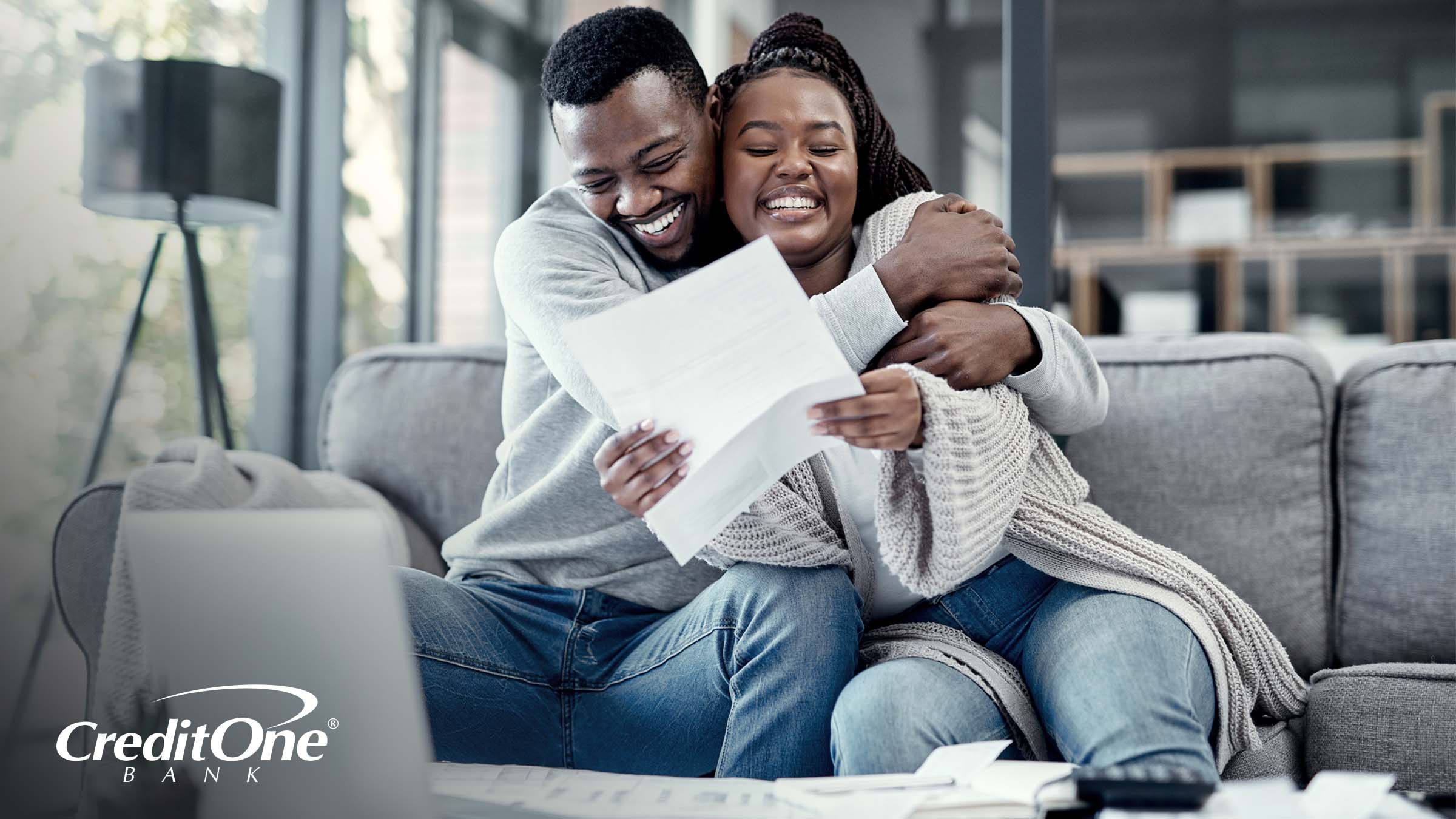 A man and a woman sit on a sofa, while a laptop and paperwork lie on a coffee table. The man happily embraces the woman, who is holding up some paperwork, as if celebrating that they paid off some credit card debt.