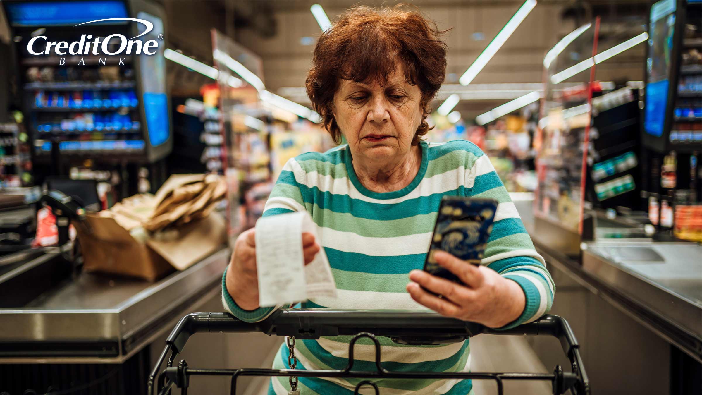 A senior woman checks her phone and receipt as she pushes her grocery cart past the store’s checkout counter with a concerned look. She may be worried about her current debt and the rising cost of groceries.
