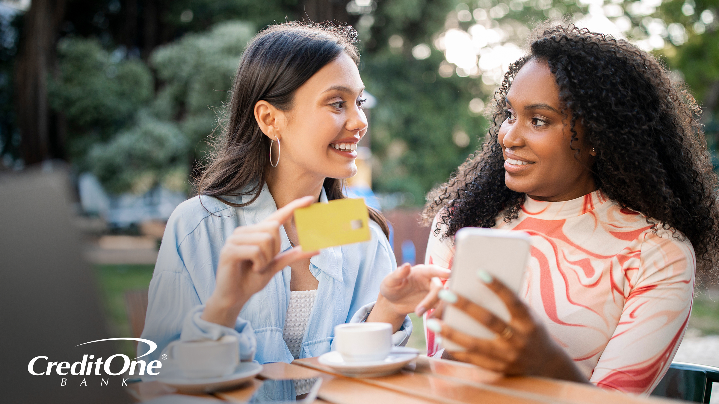 Two women are socializing at an outdoor picnic table, which has coffee mugs and a laptop on it. One woman holds up her credit card as if demonstrating the CVV number before she makes an online purchase.