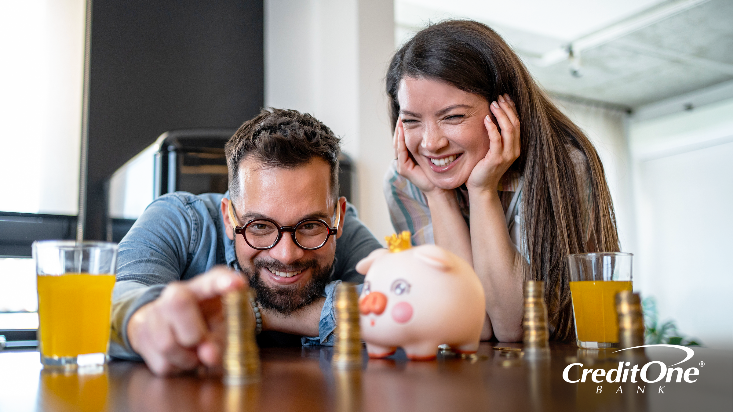 A happy couple is playing with stacks of coins on the table beside a piggy bank, separating their savings into categories like you would with both a high-yield and a traditional savings account.
