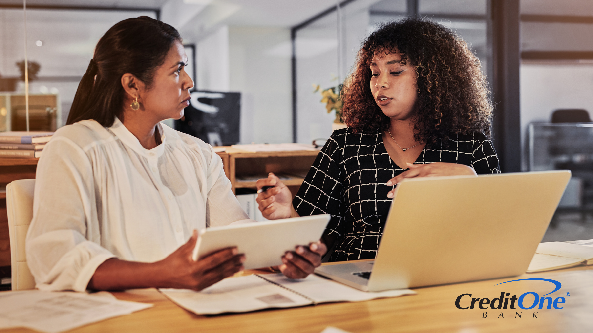 Two women have a discussion while sitting at a table. One has a laptop open while speaking and the other is listening carefully while holding on to her tablet. They may be discussing the differences between traditional and AI-based financial advice.