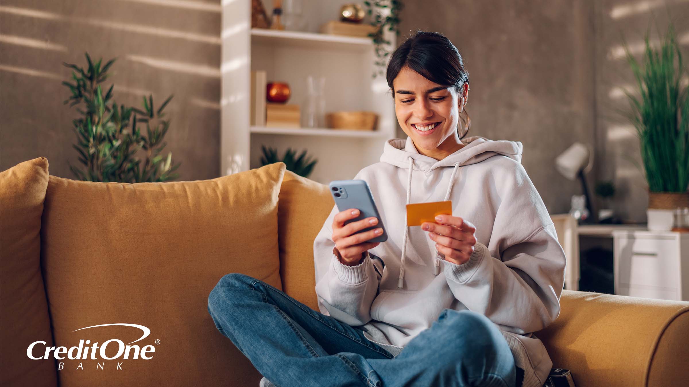 A young woman smiles as she sits on her couch with mobile phone in one hand and credit card in the other.