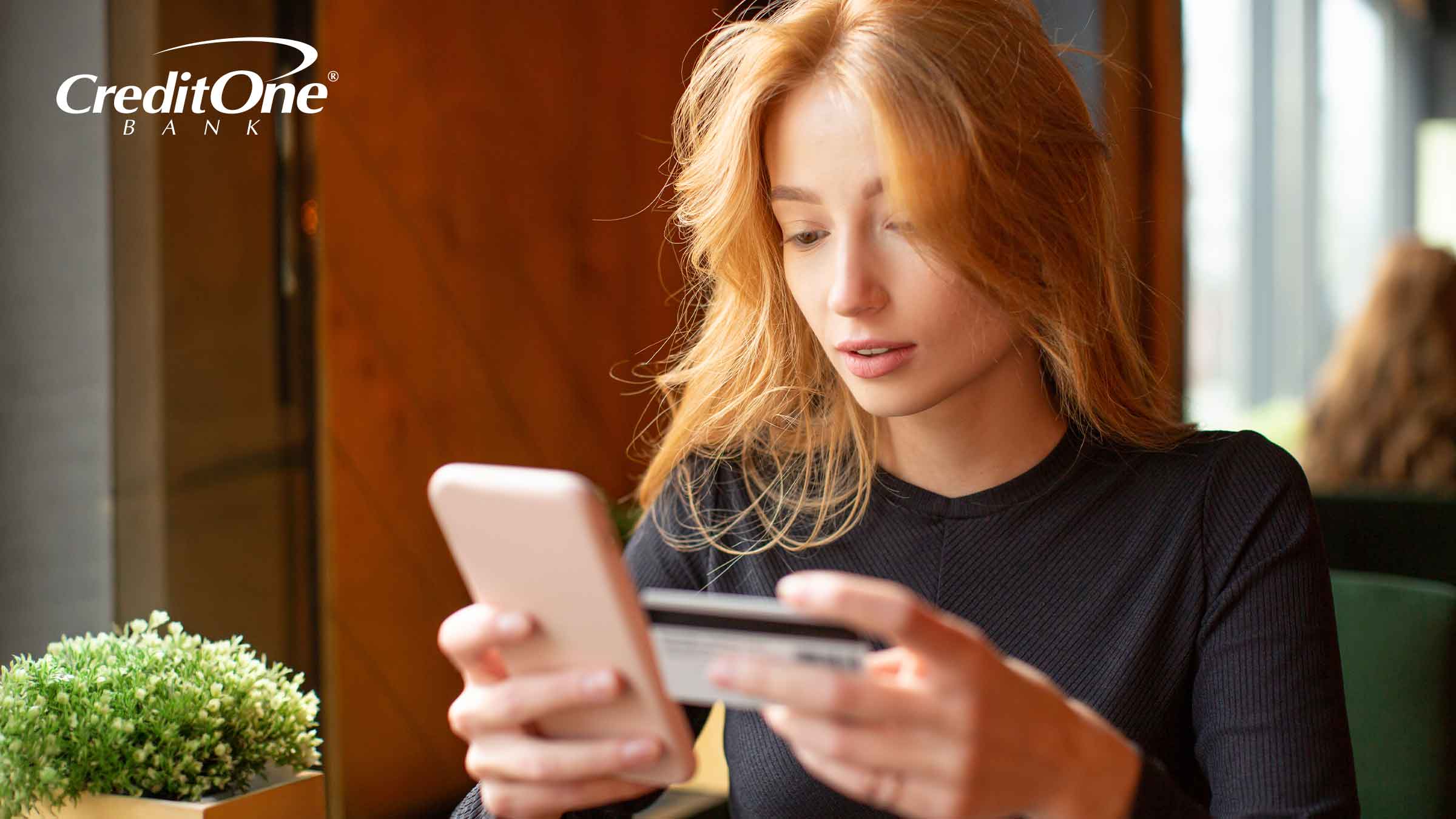 A young woman sits at a café table holding her phone in one hand and credit card in the other, perhaps taking care of making her minimum credit card payment on the go.