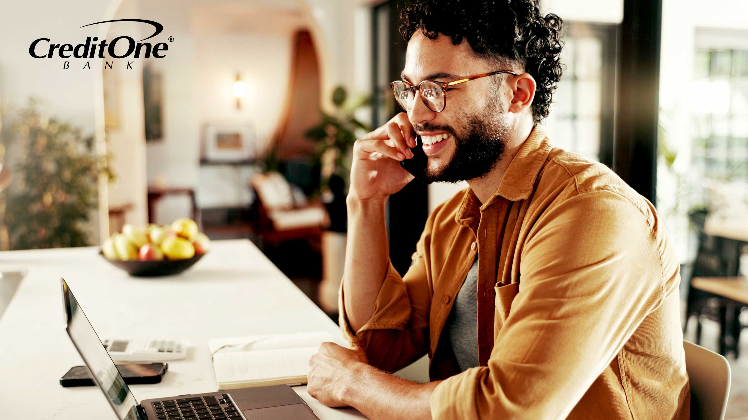 A young man sitting at a counter talks on his phone while smiling as if he’s getting good news, like an update to his credit score or loan application.