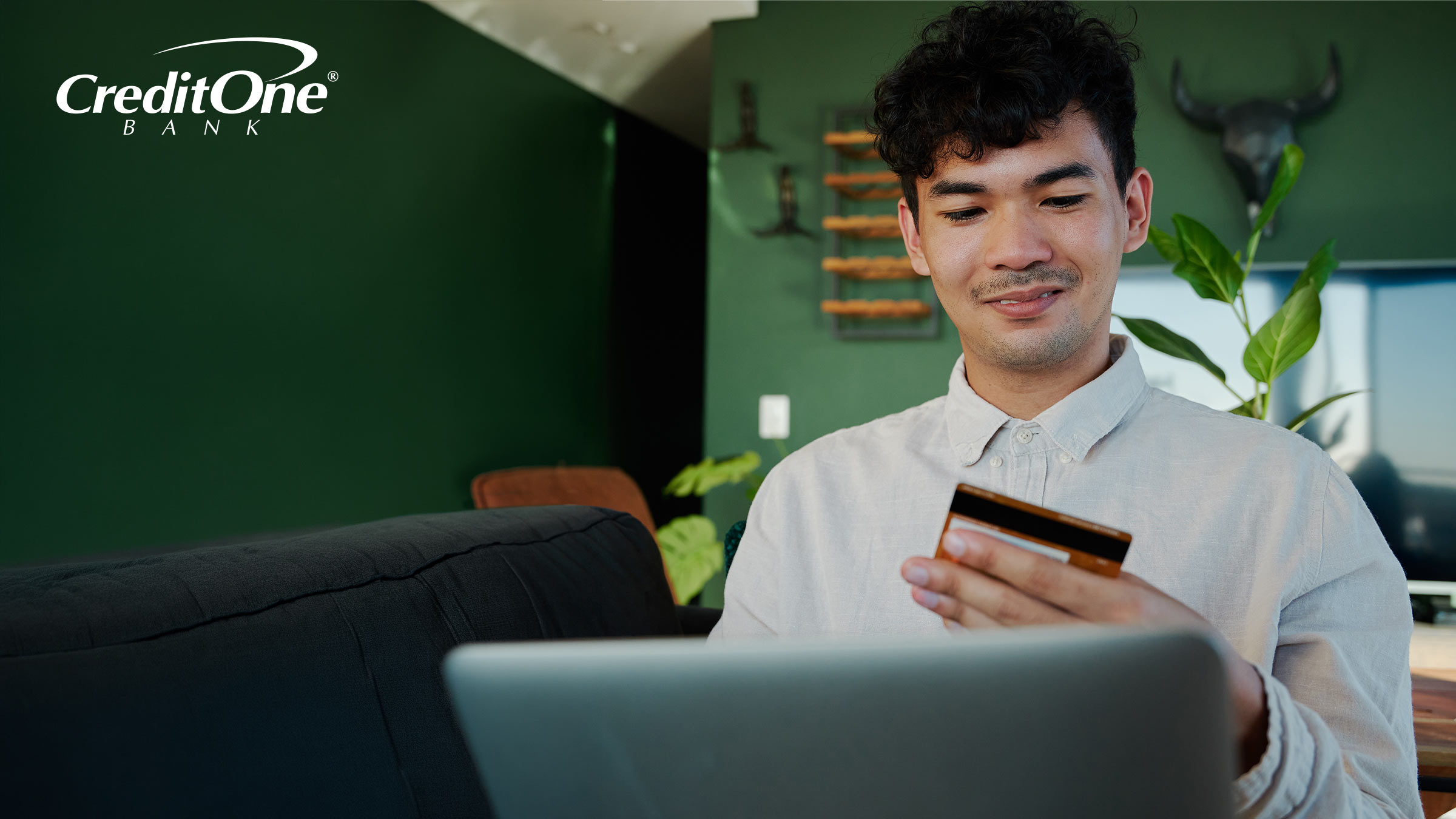A man sits at his laptop while holding his credit card in one hand, looking at it as if he’s considering whether the benefits outweigh the annual fee.