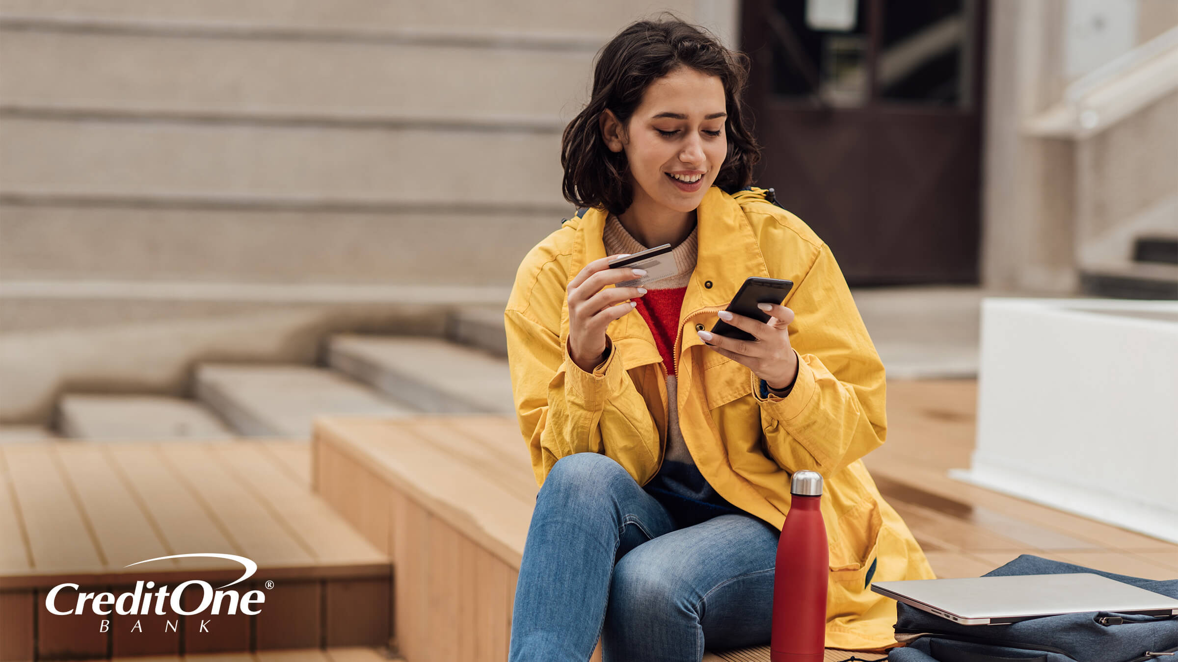 A smiling young woman sits on the steps with her backpack, laptop, and water bottle beside her. She holds a credit card in one hand and her phone in the other as if she’s monitoring her accounts while building credit.