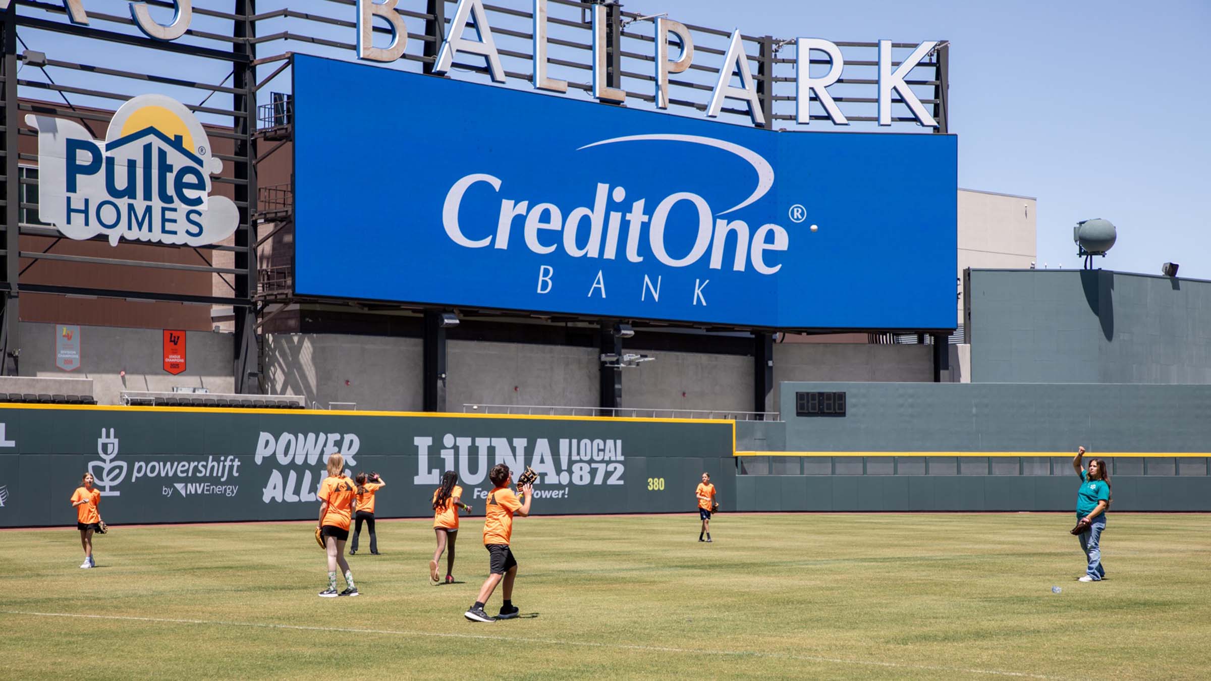YMCA students playing baseball on the Las Vegas Ballpark field with Credit One Bank staff and the Credit One Bank logo on a billboard