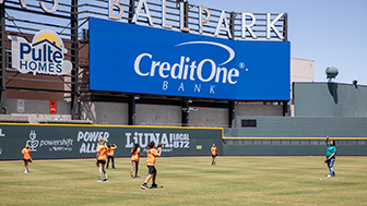 YMCA students playing baseball on the Las Vegas Ballpark field with Credit One Bank staff and the Credit One Bank logo on a billboard